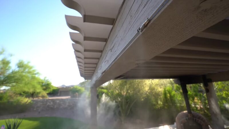 A close-up view of a misting system installed under a wooden patio pergola, releasing a fine mist to cool down the outdoor space in a lush backyard