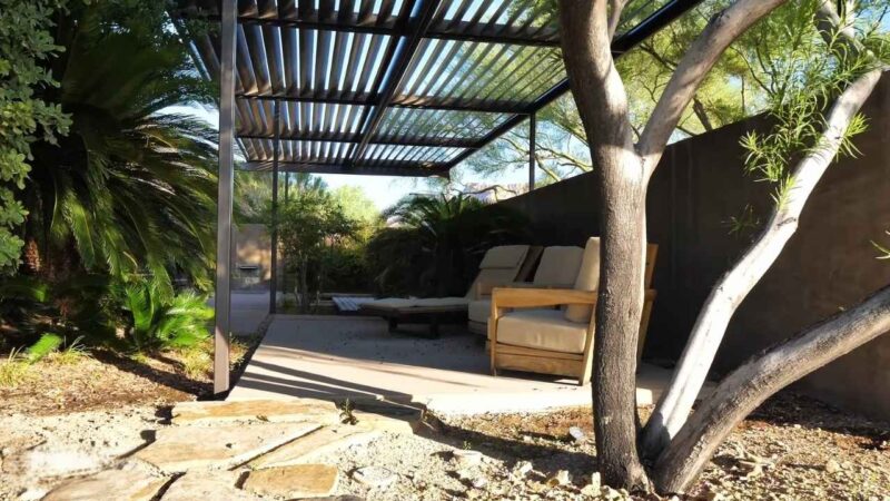 A modern shaded outdoor seating area with adjustable metal louvers, surrounded by trees and plants, creating a relaxing space with airflow and dust protection